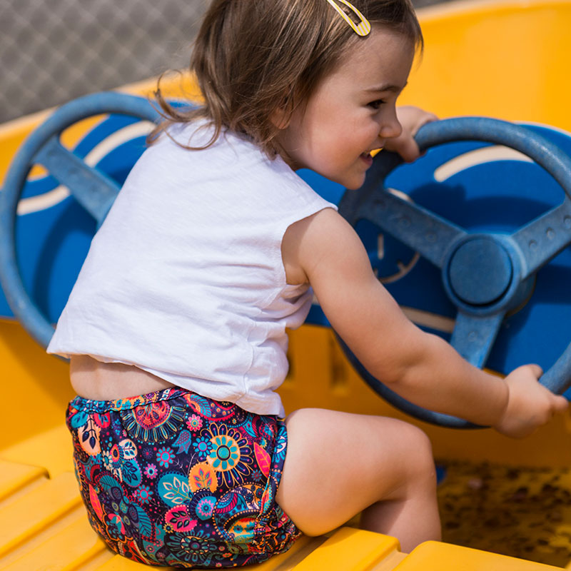 A little girl on a play boat is wearing a botanic baby diaper cover bright colorful purple, orange, pink, blue.  The diaper cover is paired with a white t-shirt.